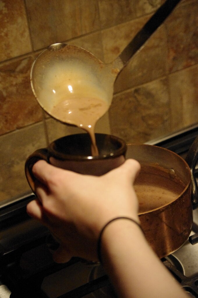 One hand is holding a mug next to a copper pot and another hand is pouring hot chocolate from a ladle into the mug