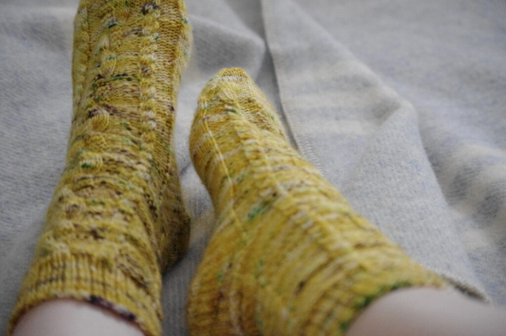 A person's feet wearing yellow knitted socks on a light blue wool blanket. 