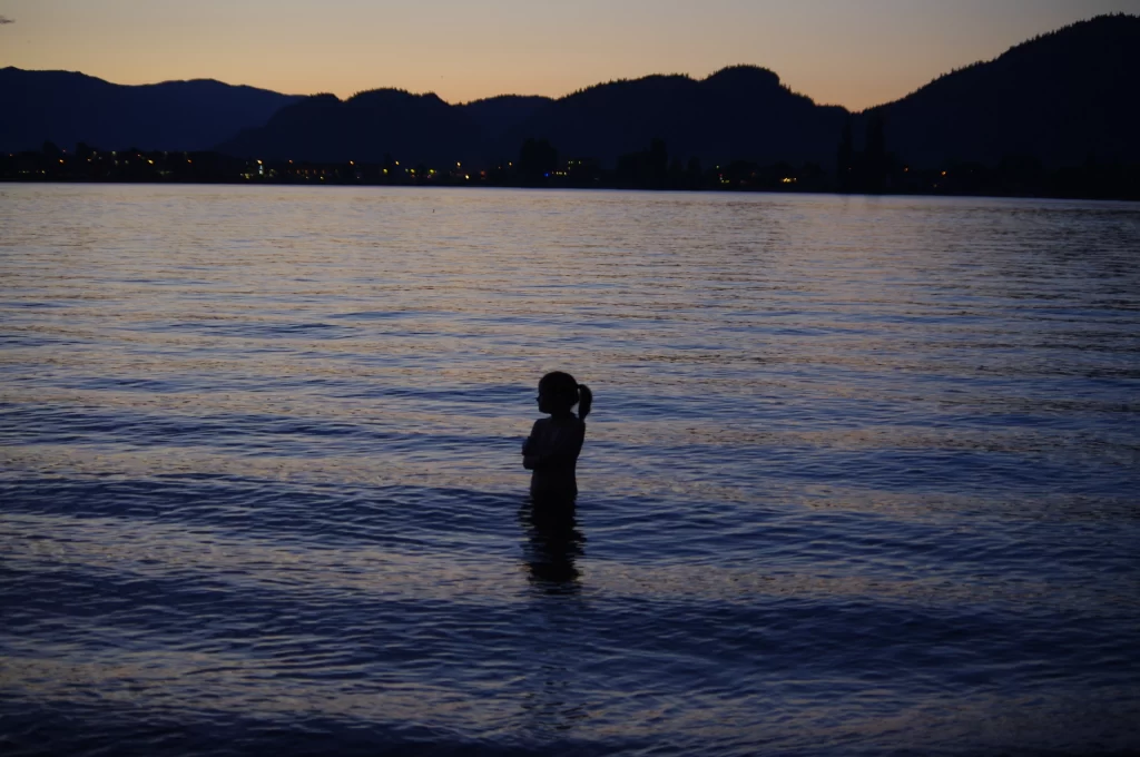 The silhouette of a girl swimming in a lake at sunset with mountains in the background